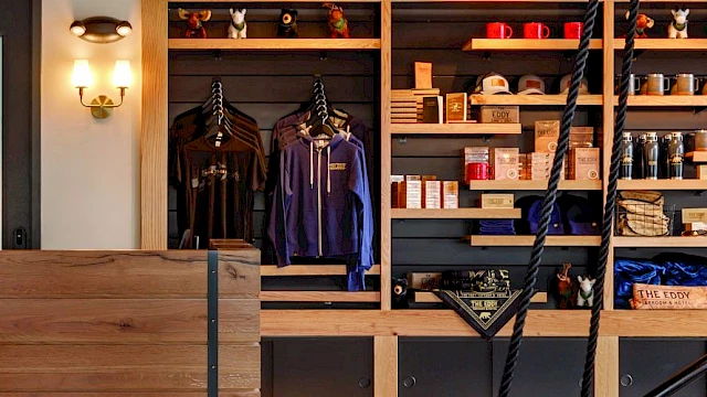 The image shows a cozy retail space with wooden shelves displaying clothing, mugs, and various items, alongside stylish wall lighting.