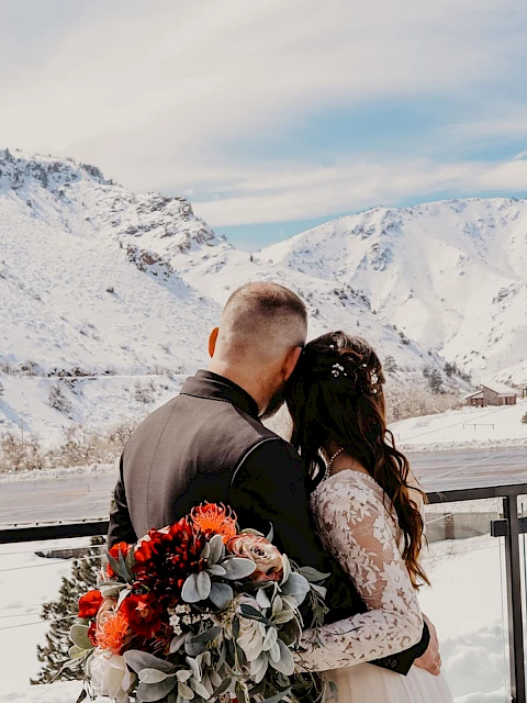 A couple is standing on a balcony, embracing with snowy mountains in the background, and one is holding a bouquet of flowers.