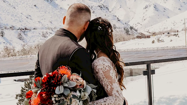 A couple is standing on a balcony, embracing with snowy mountains in the background, and one is holding a bouquet of flowers.