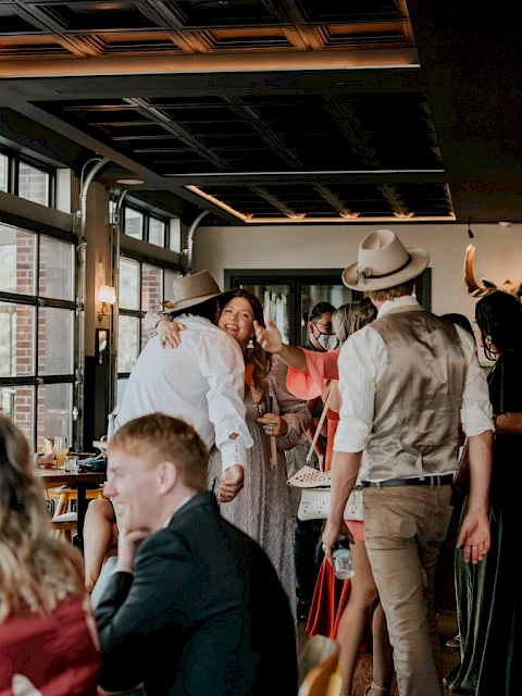 People in a cozy restaurant, some embracing and chatting, others sitting at tables. The atmosphere is warm and lively.
