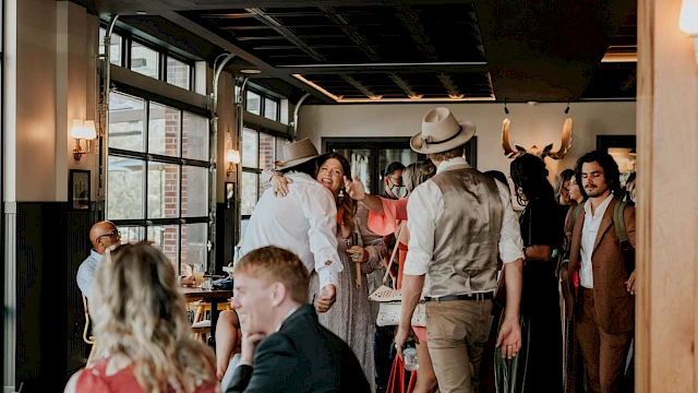 People in a cozy restaurant, some embracing and chatting, others sitting at tables. The atmosphere is warm and lively.