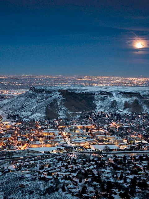 A snowy cityscape at night with a glowing full moon above, hills in the background, and city lights illuminating the buildings below.