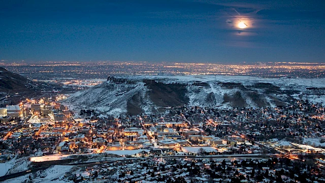 A snowy cityscape at night with a glowing full moon above, hills in the background, and city lights illuminating the buildings below.