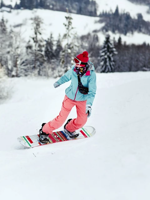 A person is snowboarding down a snowy slope wearing a blue jacket, pink pants, and a red beanie, surrounded by snow-covered trees.
