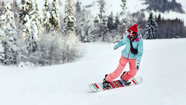 A person is snowboarding down a snowy slope wearing a blue jacket, pink pants, and a red beanie, surrounded by snow-covered trees.