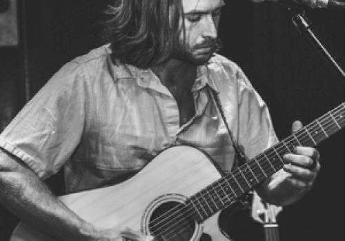A person is playing an acoustic guitar on stage, standing near a microphone, captured in a black and white photograph.