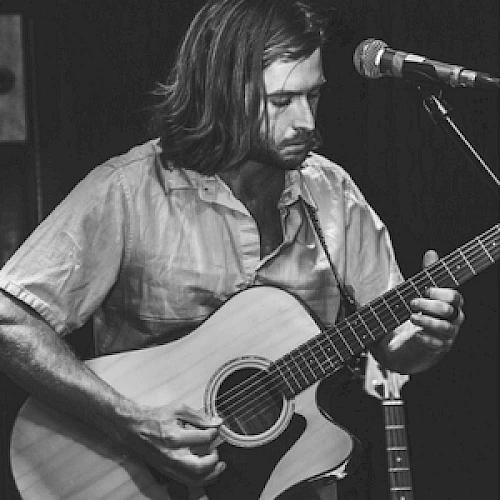 A person is playing an acoustic guitar on stage, standing near a microphone, captured in a black and white photograph.