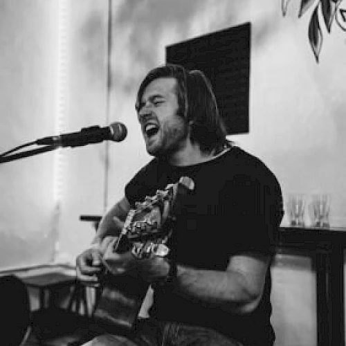 A person is passionately singing and playing a guitar into a microphone in a relaxed indoor setting.