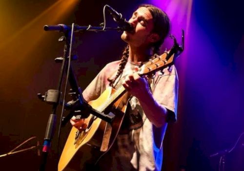 A musician is playing guitar and singing on stage under colorful lights, creating a lively and engaging performance atmosphere.