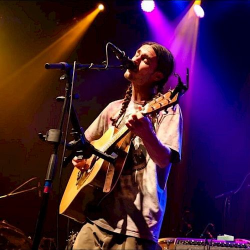 A musician is playing guitar and singing on stage under colorful lights, creating a lively and engaging performance atmosphere.