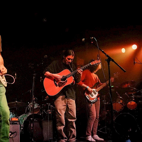 A band performs live on stage, with guitarists and a drummer under warm lighting.