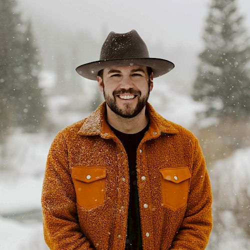 A person in a hat and orange jacket stands in a snowy landscape with trees in the background, smiling at the camera.