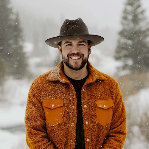 A person in a hat and orange jacket stands in a snowy landscape with trees in the background, smiling at the camera.