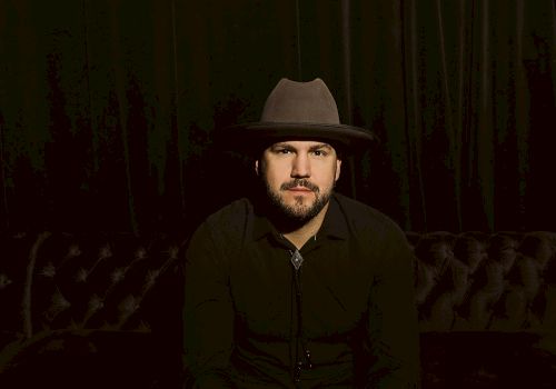 A person wearing a wide-brimmed hat and dark clothing sits on a tufted couch against a dark background.