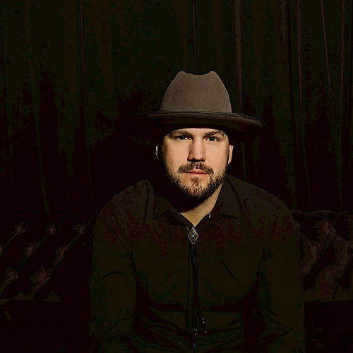 A person wearing a wide-brimmed hat and dark clothing sits on a tufted couch against a dark background.