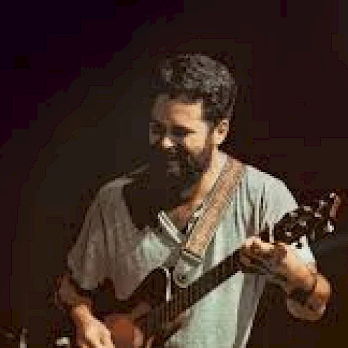 A musician with a beard is playing a guitar on stage, with a dark background and a microphone nearby.