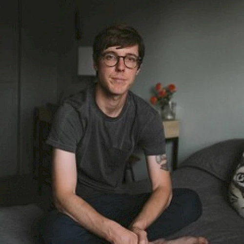 A person in glasses sits on a bed in a room with a vase of flowers behind them.