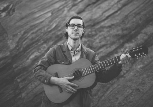 A person playing a guitar, standing against a rocky background in a black-and-white photo.