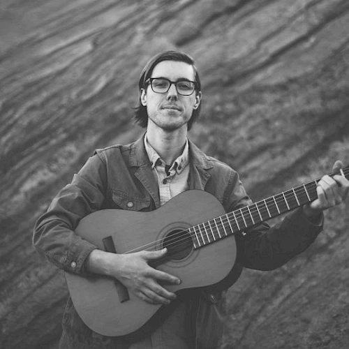 A person playing a guitar, standing against a rocky background in a black-and-white photo.