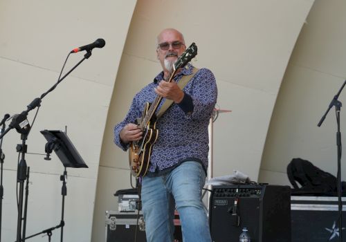 A person is playing guitar on stage near microphones and audio equipment, smiling while performing.