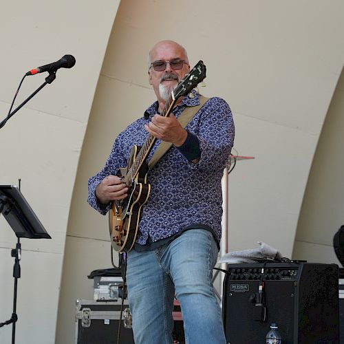 A person is playing guitar on stage near microphones and audio equipment, smiling while performing.