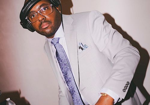 A DJ in a suit and headphones is standing at a DJ mixer, adjusting controls.