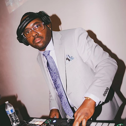 A DJ in a suit and headphones is standing at a DJ mixer, adjusting controls.