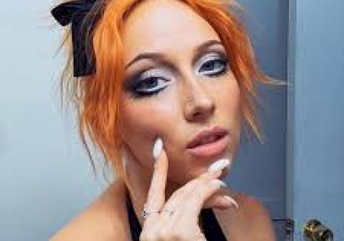 A person with orange hair styled messily, wearing dramatic eye makeup and a black bow, posing with fingers near the face indoors.