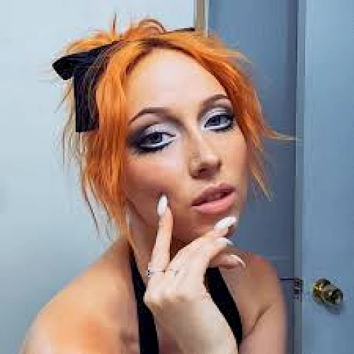 A person with orange hair styled messily, wearing dramatic eye makeup and a black bow, posing with fingers near the face indoors.