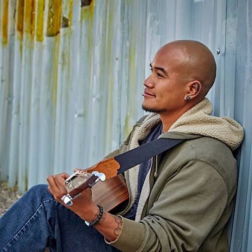 A person is sitting against a metal wall, holding a guitar and looking off to the side, with a calm expression.