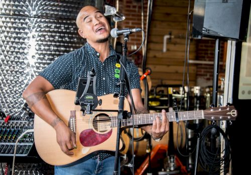 A person is passionately singing and playing an acoustic guitar, standing in front of a microphone in a music studio or similar setting.