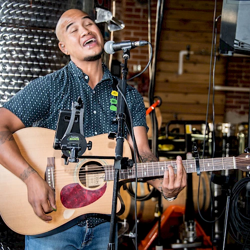 A person is passionately singing and playing an acoustic guitar, standing in front of a microphone in a music studio or similar setting.