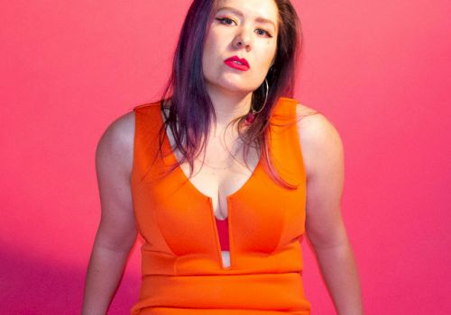 A person in an orange dress sits against a pink background, looking at the camera with arms resting on the surface.