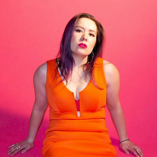 A person in an orange dress sits against a pink background, looking at the camera with arms resting on the surface.
