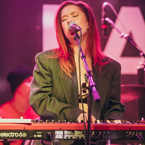 A person is passionately singing and playing a keyboard on stage, wearing a dark blazer and with a microphone in front of them.