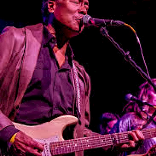 A musician is playing an electric guitar and singing into a microphone on stage, dressed in a blazer under purple lighting.
