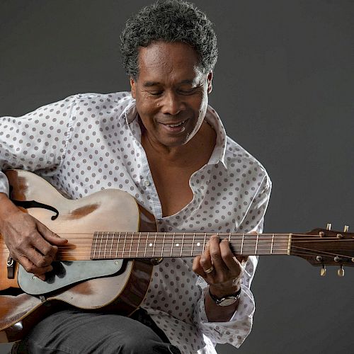 A person in a patterned shirt plays an acoustic guitar with a focused expression.
