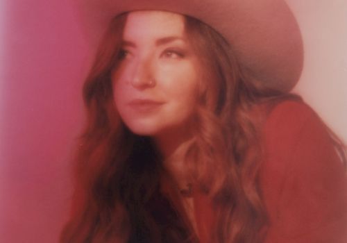 A person wearing a cowboy hat and red clothing is lying down against a softly lit, pink background, looking off to the side thoughtfully.