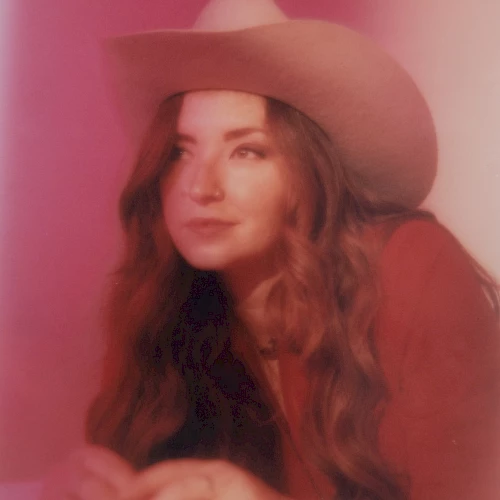 A person wearing a cowboy hat and red clothing is lying down against a softly lit, pink background, looking off to the side thoughtfully.
