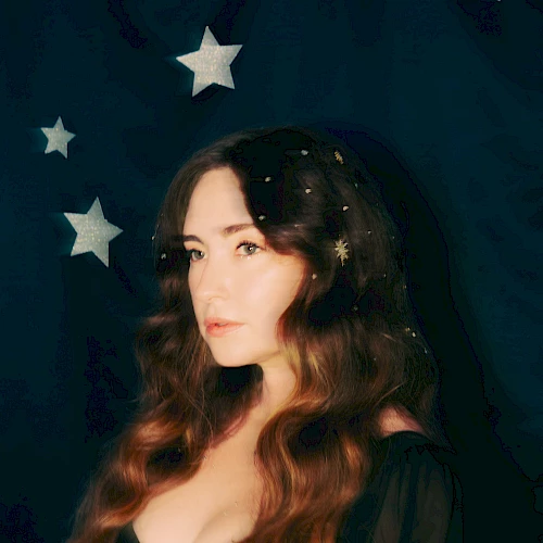 A person with long hair stands against a dark starry backdrop, wearing a black outfit with small stars in their hair.