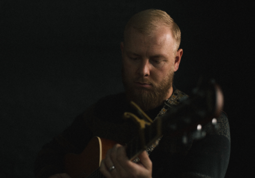 A person plays an acoustic guitar in a dimly lit setting, focusing intently on the instrument.