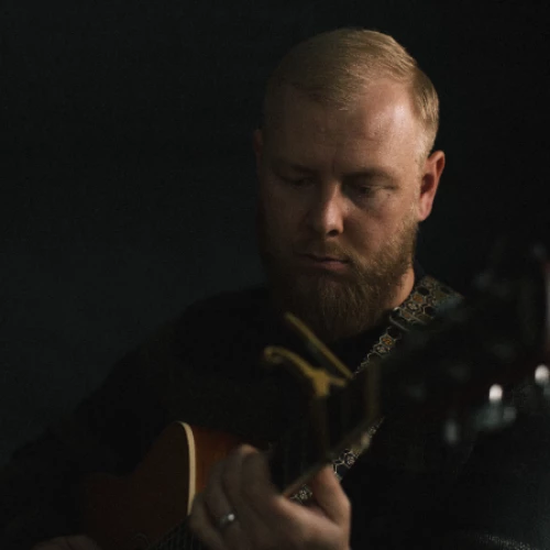 A person plays an acoustic guitar in a dimly lit setting, focusing intently on the instrument.