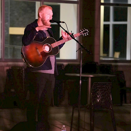 A person is playing an acoustic guitar and singing into a microphone on a dimly lit stage.