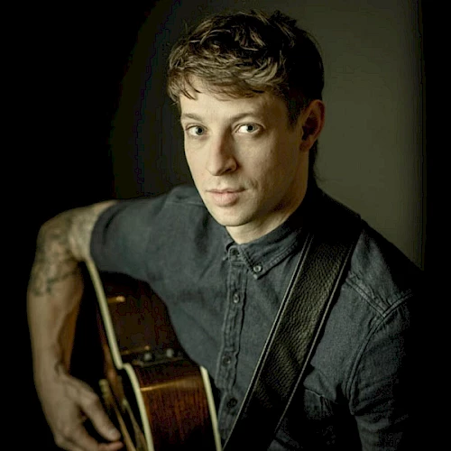 A person holding a guitar, wearing a dark button-up shirt, looking towards the camera.