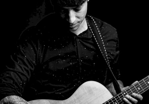 A person plays an acoustic guitar, focusing intently on the instrument, in a black and white setting.