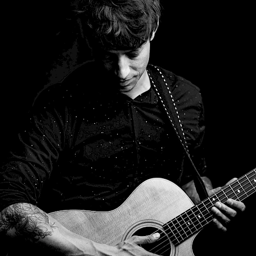 A person plays an acoustic guitar, focusing intently on the instrument, in a black and white setting.