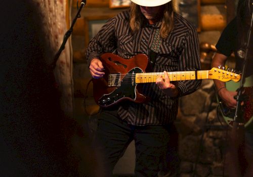 A musician wearing a hat plays an electric guitar in a dimly lit setting, possibly a live performance in a rustic venue.