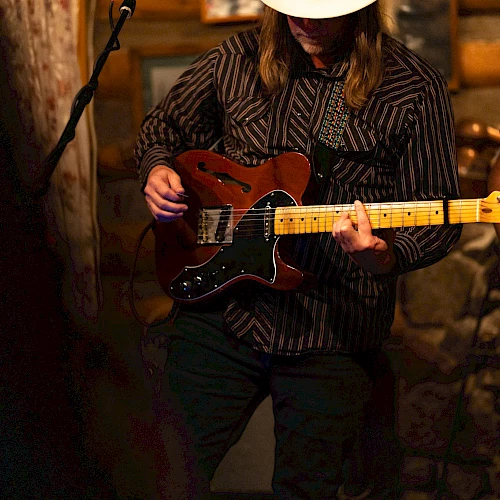A musician wearing a hat plays an electric guitar in a dimly lit setting, possibly a live performance in a rustic venue.