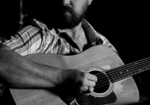 The image shows a person with a beard playing an acoustic guitar, wearing a plaid shirt.