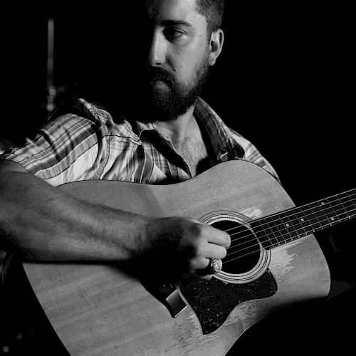 The image shows a person with a beard playing an acoustic guitar, wearing a plaid shirt.